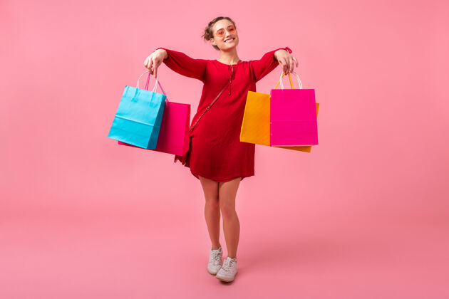 欢呼迷人的快乐微笑的时尚女性购物狂穿着红色的新潮连衣裙拿着五颜六色的购物袋在粉色的墙上孤立着 销售兴奋 时尚潮流购买长度购买