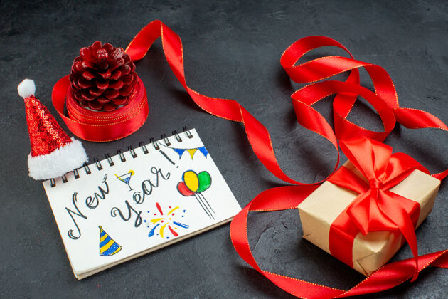 圣诞老人俯瞰一个礼物针叶树锥与红丝带和笔记本与新年写作和圣诞老人帽子美丽的礼物在黑暗的背景圣诞节鲜花文字