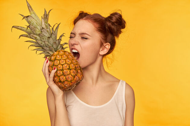 面部年轻的女士 漂亮的姜黄色的女人 带着两个小面包穿着白衬衫 闭着眼睛咬菠萝 健康的生活方式站在黄色的墙上闭嘴眼睛菠萝