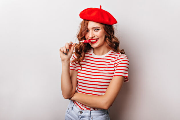 放松情绪化的法国女孩穿着t恤吃棒棒糖迷人的卷发女人享受糖果贝雷帽肖像糖果