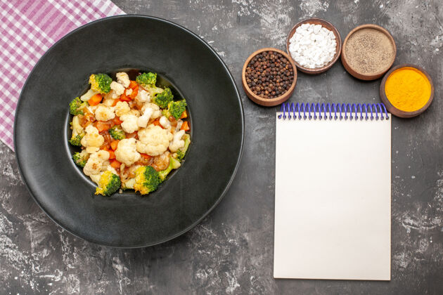 花椰菜和花椰菜顶视图西兰花和花椰菜沙拉在黑碗粉和白方格餐巾不同的香料一个笔记本在黑暗的表面煎锅笔记本碗