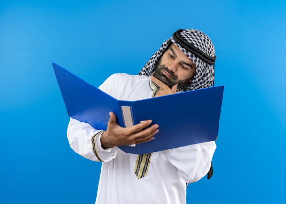 站身着传统服饰的阿拉伯商人拿着打开的文件夹严肃地看着它站在蓝色的墙上穿认真持有