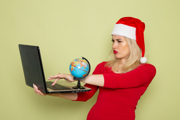 漂亮正面图美丽的女性手持小地球仪和笔记本电脑在绿墙上圣诞雪假日新年感慨地球仪视图雪