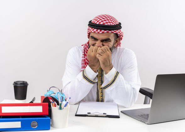 电脑身着传统服装的阿拉伯商人坐在桌旁 手里拿着笔记本电脑 紧张地咬着指甲在办公室工作笔记本电脑咬阿拉伯语
