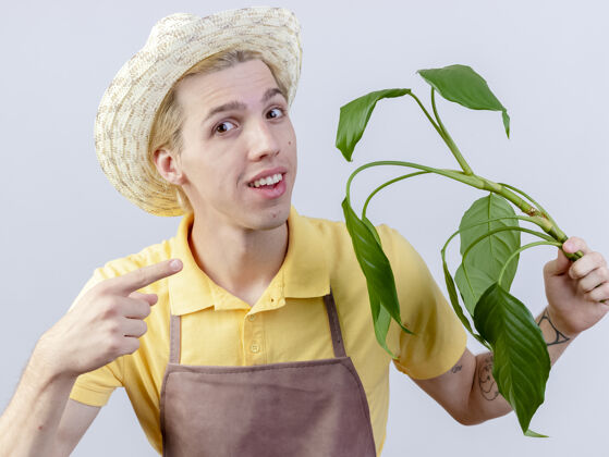 连身衣年轻的园丁 穿着连体衣 戴着帽子 拿着一株植物 用食指指着它 站在白色的背景上 满脸笑容目录脸穿