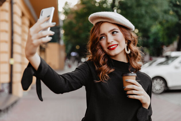 散步快乐的红发女孩捧着咖啡在户外摆姿势无忧无虑的法国小姐在街上自拍帽子手机肖像