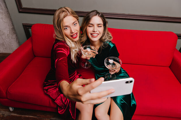 手机一个穿红衣服的金发女人和朋友一起喝香槟一个穿绿衣服的女人在沙发上摆姿势朋友室内性感