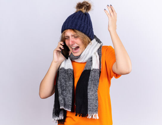 感觉不健康的年轻女子穿着橙色t恤 脖子上围着暖和的围巾 戴着帽子 感觉很糟糕 患流感 在手机上说话 表情咄咄逼人 在白墙上担心不健康流感年轻