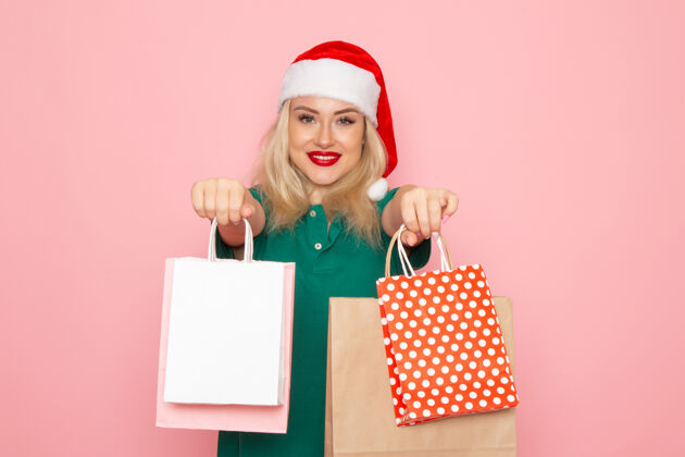 购物者正面图年轻女性手持圣诞礼物在粉红色墙上的照片模型新年假期包年轻女性观赏圣诞节