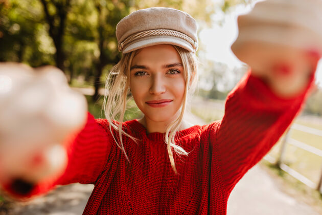 闪光可爱的金发女孩戴着漂亮的时髦帽子 穿着红色套头衫在秋季公园自拍漂亮年轻女人