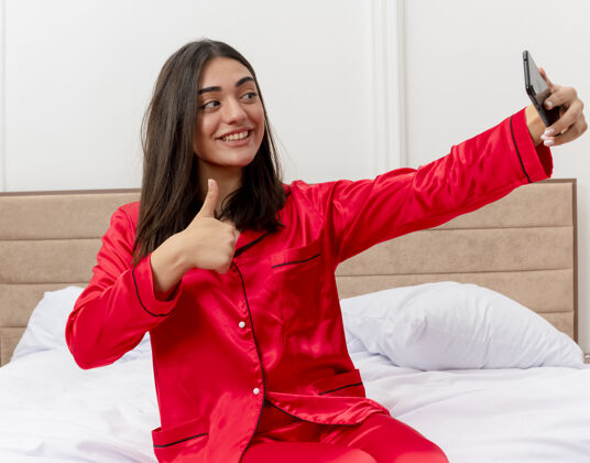 女士穿着红色睡衣的年轻漂亮女人坐在床上用智能手机自拍 微笑着在卧室内部灯光背景下竖起大拇指自拍红色睡衣