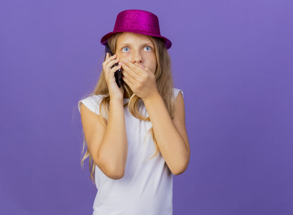 漂亮戴着节日礼帽的漂亮小女孩在用手机聊天时被震惊了 生日派对的概念站在紫色的背景上手机站着女孩