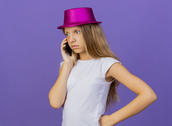 说话戴着节日礼帽的漂亮小女孩在手机上乱说 生日聚会的概念站在紫色的背景上女孩电话站