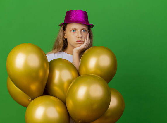 生日戴着节日礼帽的漂亮小女孩和一群巴隆人困惑地看着一边 绿色背景下站着生日派对的概念绿色站女孩