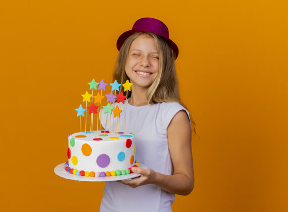 生日戴着节日礼帽的漂亮小女孩手拿生日蛋糕 兴奋地站在橙色背景上 生日派对的概念橙色节日女孩