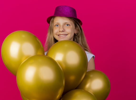 束戴着节日礼帽的快乐可爱的小女孩和一群巴隆人看着镜头笑脸盈盈 粉色背景下站着生日派对的概念站帽子快乐
