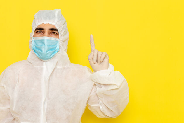 男性身穿白色特殊防护服 带无菌口罩的男性科学工作者正面图抬起手指放在黄色办公桌上科学工作者化学彩色危险照片医学化学工人