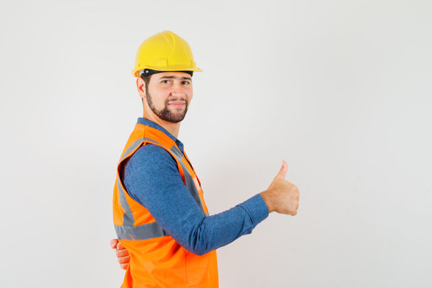 建筑年轻的建筑工人穿着衬衫 背心 头盔 竖起大拇指 看上去很高兴设备建筑工人工厂