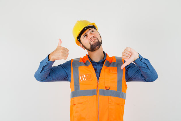 工人年轻的建筑工人穿着衬衫 背心 头盔上下竖起大拇指 看上去犹豫不决建筑衬衫工具