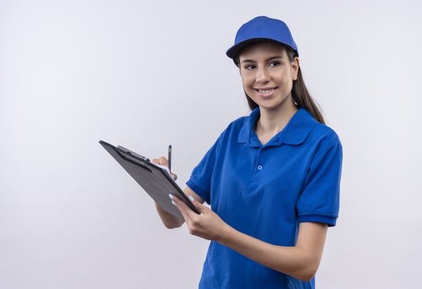 剪贴板穿着蓝色制服 戴着帽子 拿着写字板和钢笔 脸上带着自信的微笑 年轻的送货员站在一旁看着放在一边脸年轻
