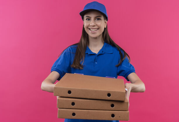 年轻身穿蓝色制服 戴着帽子 手里拿着一叠比萨饼盒的年轻送货女孩自信地微笑着看着镜头站着披萨看着