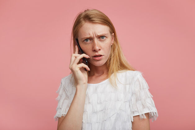 手机困惑的年轻红发女子身穿白色节日t恤 站在粉色背景下 不愉快的电话交谈 困惑地看着镜头站立女性雀斑