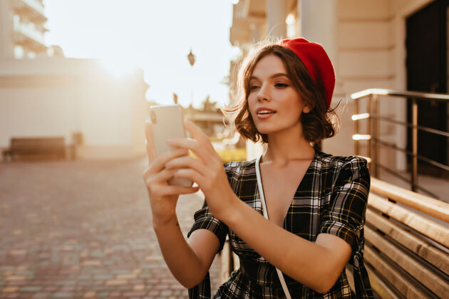 配饰可爱的黑发女士在阳光明媚的日子里为自己拍照穿着时尚的法国贝雷帽手持智能手机的快乐女模特的肖像红色贝雷帽法国自拍