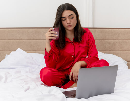床穿着红色睡衣的年轻漂亮女人坐在床上拿着一杯咖啡在笔记本电脑上工作 在灯光背景下严肃地看着卧室内部的屏幕严肃笔记本电脑坐着