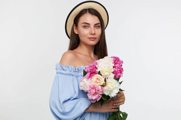 年轻十几岁的女孩 快乐的女人 深色长发 戴着帽子 穿着蓝色漂亮的裙子 手里拿着漂亮的花 隔着白色的墙看着孤零零的人室内头发成人