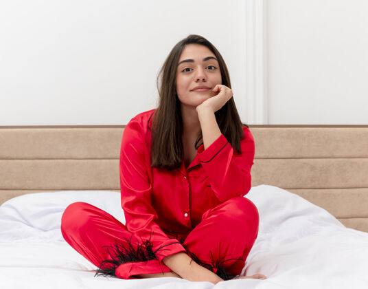 漂亮穿着红色睡衣的年轻漂亮女人坐在床上 头靠在手上看着相机 在灯光的背景下 在家里微笑室内红色手
