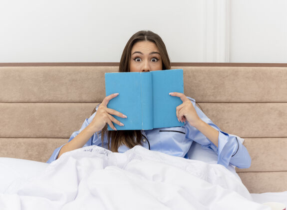 看着穿着蓝色睡衣的年轻美女坐在床上拿着书看着相机惊讶地在卧室的室内灯光背景下年轻女人床