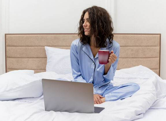 室内穿着蓝色睡衣的有趣年轻漂亮女人坐在床上喝着咖啡在笔记本电脑上工作愉快而积极地看着卧室内部的灯光背景咖啡看女士