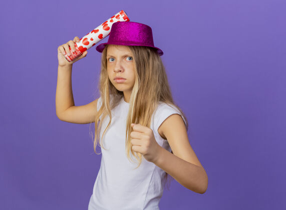 节日戴着节日礼帽的漂亮小女孩拿着派对饼干看着镜头 愤怒的脸握紧拳头 生日派对的概念站在紫色的背景上女孩站着饼干