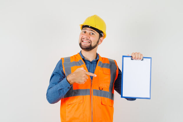 制服年轻的建筑工人穿着衬衫 背心 头盔指着剪贴板 愉快地看着 前视图工厂职业工业