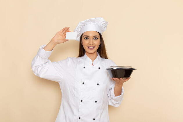 前面身穿白色厨师服的年轻女厨师正拿着黑色碗和卡片站在浅白的墙上专业年轻人膳食