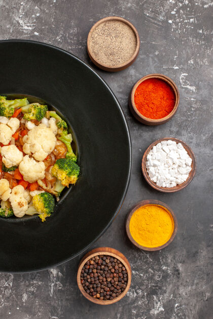 午餐俯视图西兰花和花椰菜沙拉在黑色椭圆形盘子里不同的香料在小碗里在黑暗的表面肉炊具煎锅