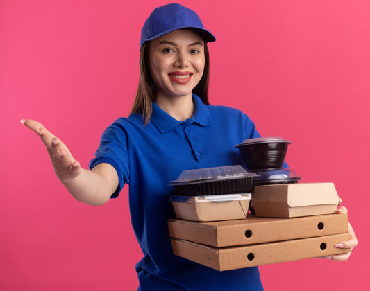 披萨面带微笑的漂亮送货员穿着制服 手拿食物包和比萨饼盒上的容器 粉红色的墙上有复印空间点漂亮制服