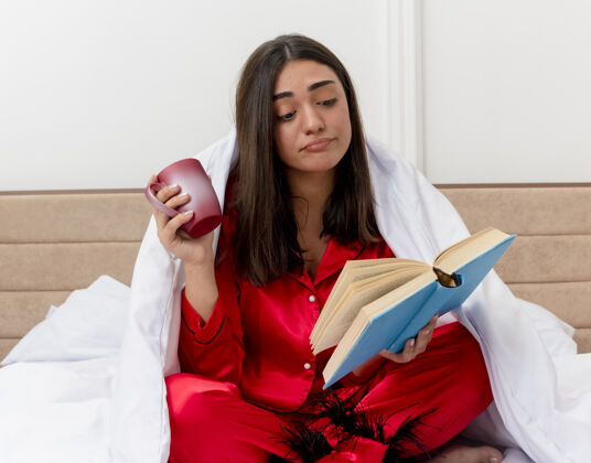 卧室穿着红色睡衣的年轻漂亮女人坐在床上 用毯子裹着一杯咖啡 在卧室的室内灯光背景下看书漂亮床毯子