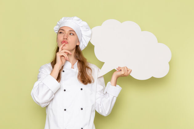 快乐身穿白色厨师服的女厨师的正面图绿色墙壁上的思考和手持白色标志女性思考厨房