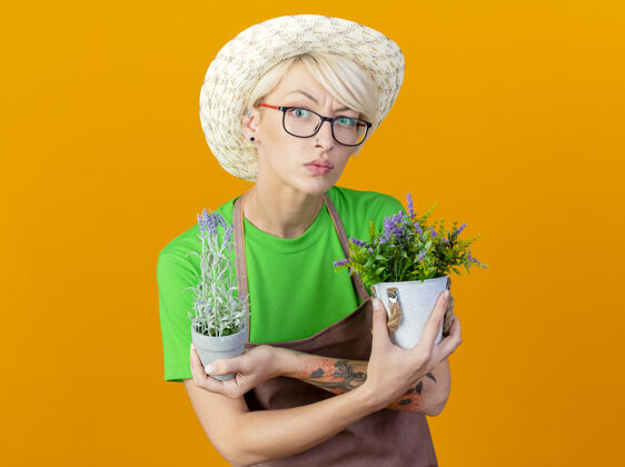 女人围裙上留着短发 戴着帽子 手里拿着盆栽植物 站在橙色背景下困惑地看着摄像机的年轻园丁女人头发立场围裙