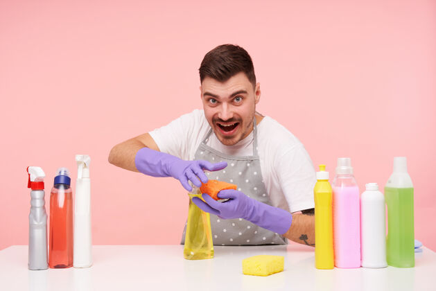 家庭一个年轻英俊的黑发男人 穿着粉红色的制服 在打扫房间的时候 把家用化学品涂在海绵上洗涤剂肖像化学品