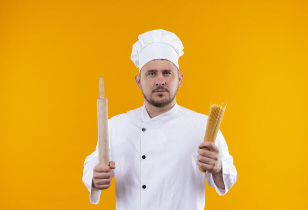 烹饪年轻帅气的厨师穿着厨师制服 手里拿着擀面杖和意大利面 看起来孤立在橙色的空间里拿着意大利面厨师