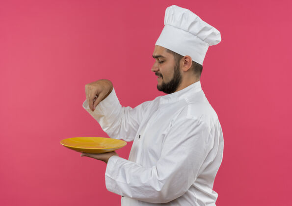 微笑面带微笑的年轻男厨师身着厨师制服 端着盘子 在粉红色的空间里加盐隔离添加盘子持有