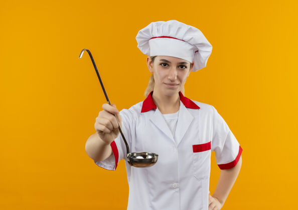 伸展年轻漂亮的厨师穿着厨师制服 手放在腰上 伸出勺子 孤立在橙色的空间里漂亮厨师制服