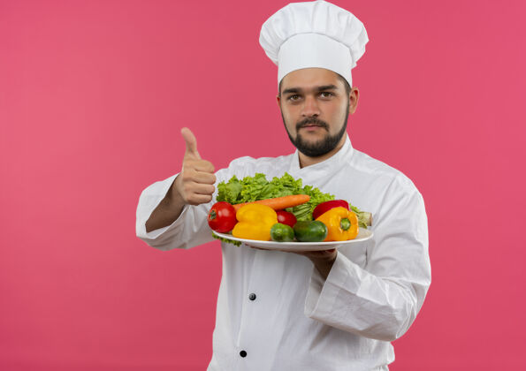 蔬菜年轻的男厨师穿着厨师制服 拿着盘子里的蔬菜 在粉红色的空间里孤立地竖起大拇指厨师盘子年轻