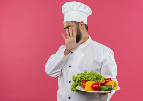 持有年轻的男厨师穿着厨师制服拿着盘子里的蔬菜 并没有孤立地在粉红色的空间里做手势手势盘子厨师