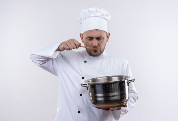 拿着年轻帅气的厨师穿着厨师制服拿着锅子 把勺子放在嘴上 隔离在空白处嘴勺子年轻