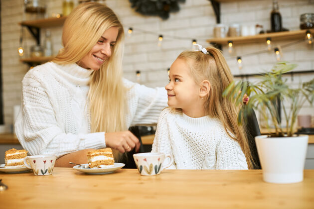 女人幸福的年轻母亲 一头金色的长发 和可爱的女儿在厨房里摆姿势 坐在餐桌旁 喝茶 吃蛋糕 面带微笑 谈笑风生童年孩子妈妈