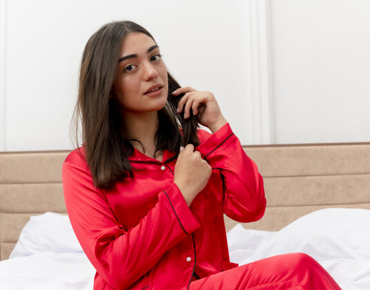坐着穿着红色睡衣的年轻美女坐在床上讲着手机 在灯光的背景下 卧室里看起来很自信红色床说话