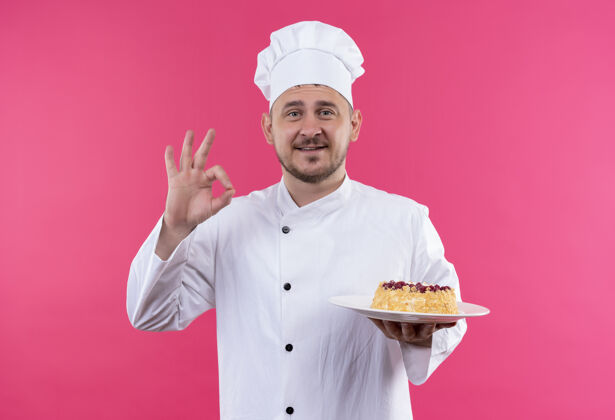 厨师笑容可掬的年轻帅哥厨师身着厨师制服拿着一盘蛋糕做ok标志孤立在粉色的空间里粉色好的帅气